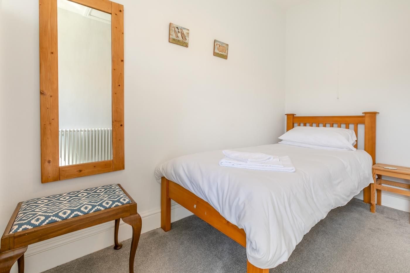 A bedroom with a single bed, a wooden nightstand, a bench with a patterned cushion, and a mirror on the wall.