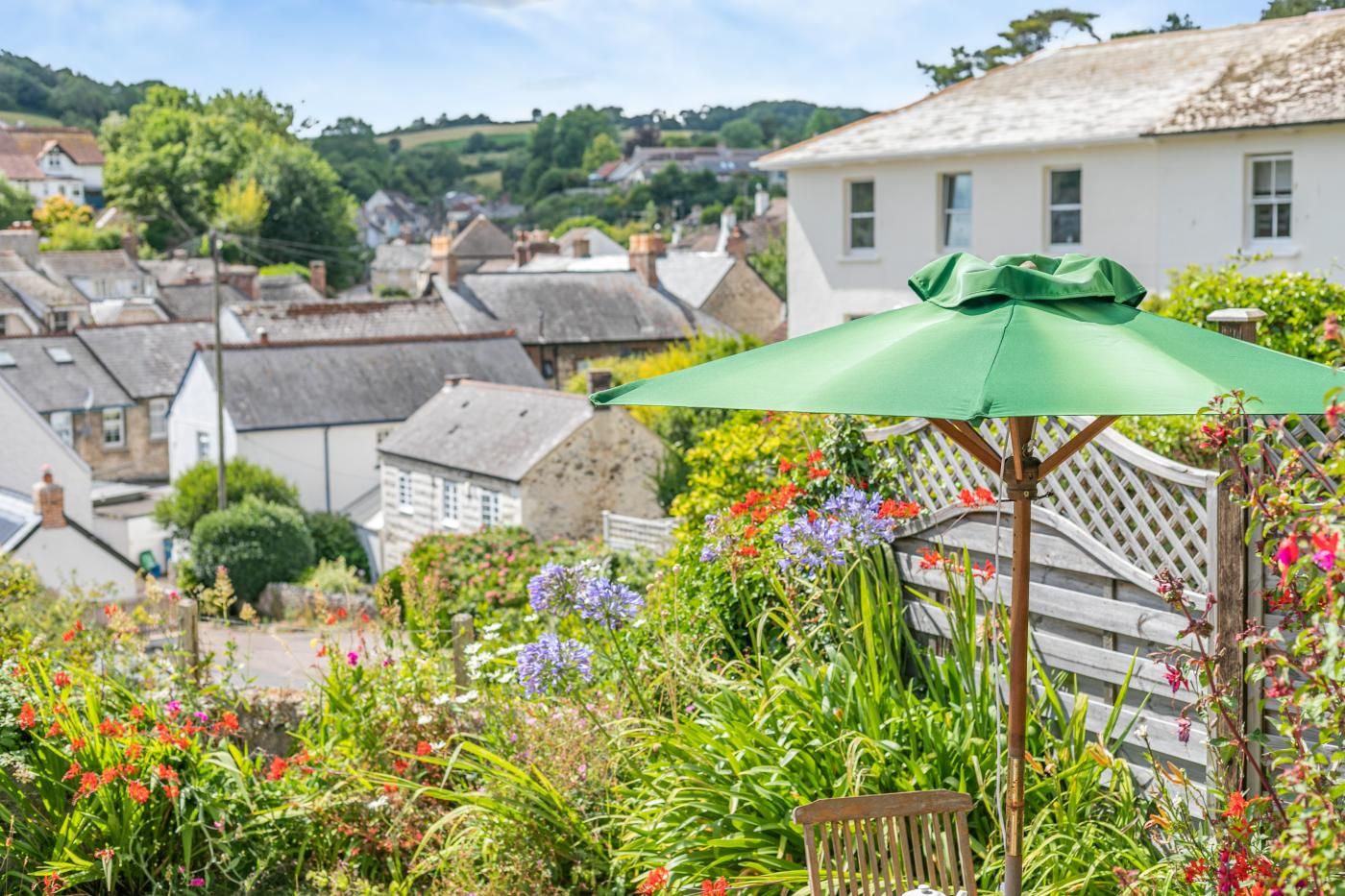 En have med en træbænk og en grøn parasol, omgivet af farverige blomster og en hvid bygning i baggrunden.