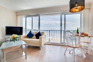 Ein modernes Wohnzimmer mit einer Couch, einem Glastisch, einem Fernseher und einem Balkon mit Blick auf den Strand.