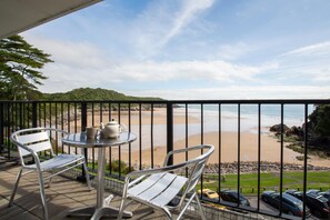 A balcony with two chairs and a table, overlooking a beach with a view of the ocean and distant hills. - 1 Bed in Caswell Bay (oc-hh9206) (Bishopston)