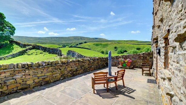 A stone-walled patio with a table and chairs, overlooking rolling hills and a stone wall. - 3 Bed in Gunnerside (oc-ds067) (Gunnerside)