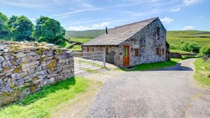 A stone house with a wooden door and a metal gate, surrounded by a stone wall and a gravel driveway. - 3 Bed in Gunnerside (oc-ds067) (Gunnerside)
