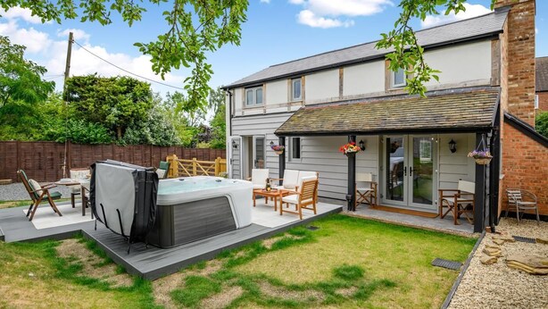 A two-story house with a patio area featuring a hot tub, outdoor seating, and a covered patio with hanging flower baskets. - 3 Bed in Alfrick (oc-s32386) (Alfrick)