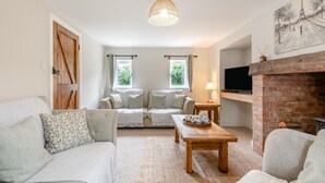 A cozy living room with a sofa, a coffee table, and a fireplace. There are two windows with curtains, a wooden door, and a painting on the wall. - 3 Bed in Alfrick (oc-s32386) (Alfrick)