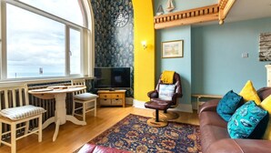 A living room with a large window, a television, a sofa with colorful cushions, a patterned rug, and a wall clock. - 2 Bed in Whitby (oc-w507) (Whitby)