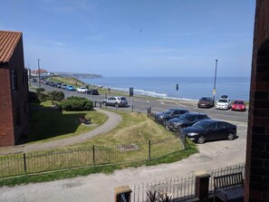 Uma estrada costeira com carros estacionados, uma calçada e vista para o mar.