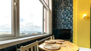 A room with a view of the sea, a round wooden table with woven placemats, a white chair, and a wall with a clock and patterned wallpaper. - 2 Bed in Whitby (oc-w507) (Whitby)
