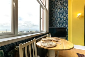 Quarto com vista para o mar, mesa de madeira redonda com jogos americanos trançados, cadeira branca e parede com relógio e papel de parede estampado.