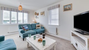 A living room with a blue sofa, a coffee table with pastries, a TV, and a radiator. - 3 Bed in Whitby (oc-w1001) (Whitby)