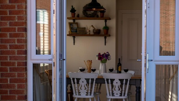 An open sliding glass door leading to a dining area with a wooden table set for two, a white chair, and a small shelf with decorative items. - 3 Bed in Whitby (oc-w1001) (Whitby)