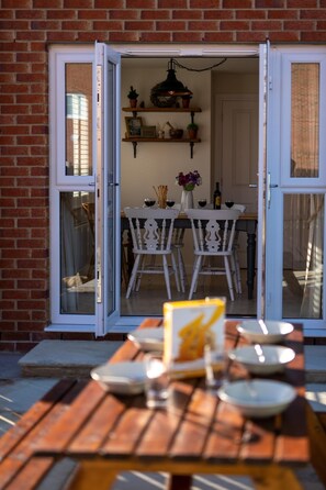 Porta finestra aperta che dà su una zona pranzo con tavolo in legno apparecchiato per due, una sedia bianca e un piccolo ripiano con oggetti decorativi.