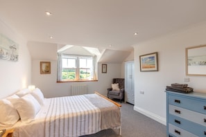 Ein Schlafzimmer mit Bett, Fenster, Sessel, Kommode und Bildern an den Wänden.
