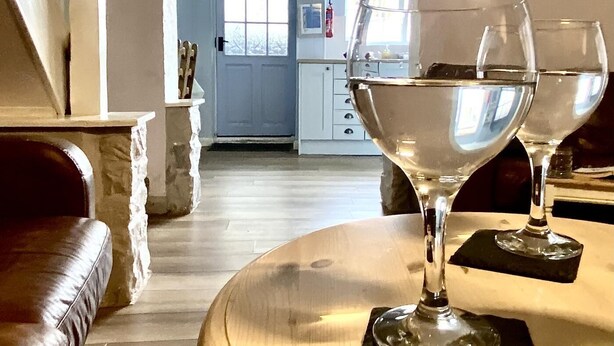 A living room with a wooden table and two wine glasses. There is a brown leather sofa and a white archway leading to a kitchen with a blue door. - 2 Bed in Whitby (oc-w985) (Whitby)