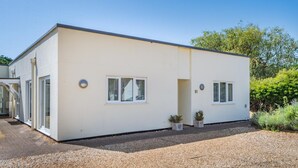A modern, single-story building with a flat roof and a white exterior. There are two windows on the front side, one on each side, and a door on the left. The building is surrounded by gravel and has a small garden area with two potted plants. - 2 Bed in Old Hunstanton (oc-s32361) (Hunstanton)