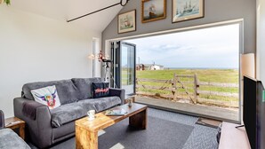 Un salon moderne avec un canapé gris, une table basse en bois et une grande fenêtre donnant sur la mer et un champ verdoyant.