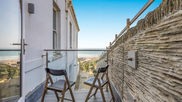 En balkon med to stole, udsigt til stranden og en stenvæg.