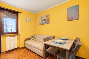 Un área de comedor con una mesa lista para dos personas, un sofá, un radiador y una ventana con cortinas estampadas.