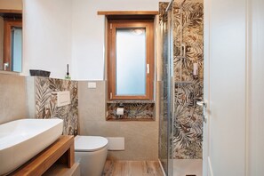 Ein Badezimmer mit einem Holzbadschränken, einer Toilette, einer Duschkabine mit Glastür und einem Fenster über dem Waschbecken.