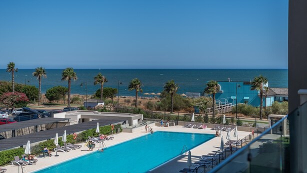 Zona de piscina con vista al mar, palmeras y un edificio de fachada azul.