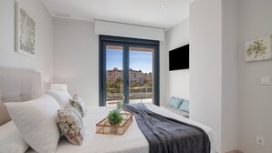 Un dormitorio moderno con una cama grande, vistas de edificios a través de un ventanal y un televisor montado en la pared.