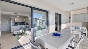 Un salón moderno con mesa de vidrio, muebles blancos y vistas a la ciudad a través de amplias ventanas.