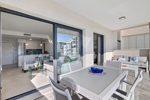 Ein modernes Wohnzimmer mit einem Glastisch, weißen Möbeln und Blick auf die Stadt durch große Fenster.