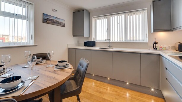 Eine moderne Küche mit Essbereich, ausgestattet mit einem Holztisch, der für eine Mahlzeit gedeckt ist, einem eingebauten Backofen und einem Fenster mit Jalousien.