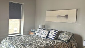 A bedroom with a large bed, a window with blinds, a painting on the wall, and a nightstand with a lamp.