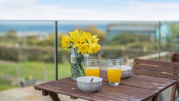 Une table en bois sur laquelle se trouve un vase de fleurs jaunes, deux verres de jus d’orange et un bol, le tout placé sur un balcon donnant sur un paysage.