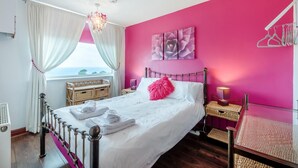 Une chambre à coucher comprenant un lit, une table de chevet, une lampe, une fenêtre avec des rideaux, une œuvre d’art accrochée au mur et une vue sur l’extérieur.