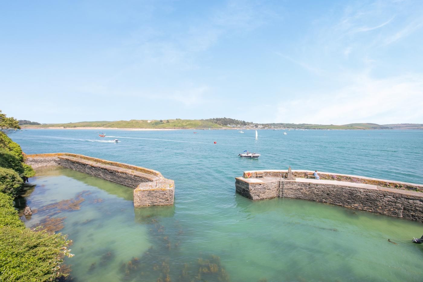 A coastal scene with a stone pier extending into calm waters, surrounded by greenery and a clear sky.