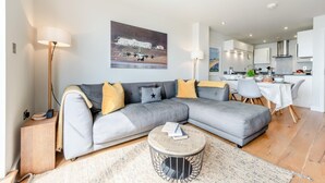 A modern living room with a grey sectional sofa, a round coffee table, and a kitchen in the background. - 2 Bed in Saunton (oc-10ocea) (Croyde)