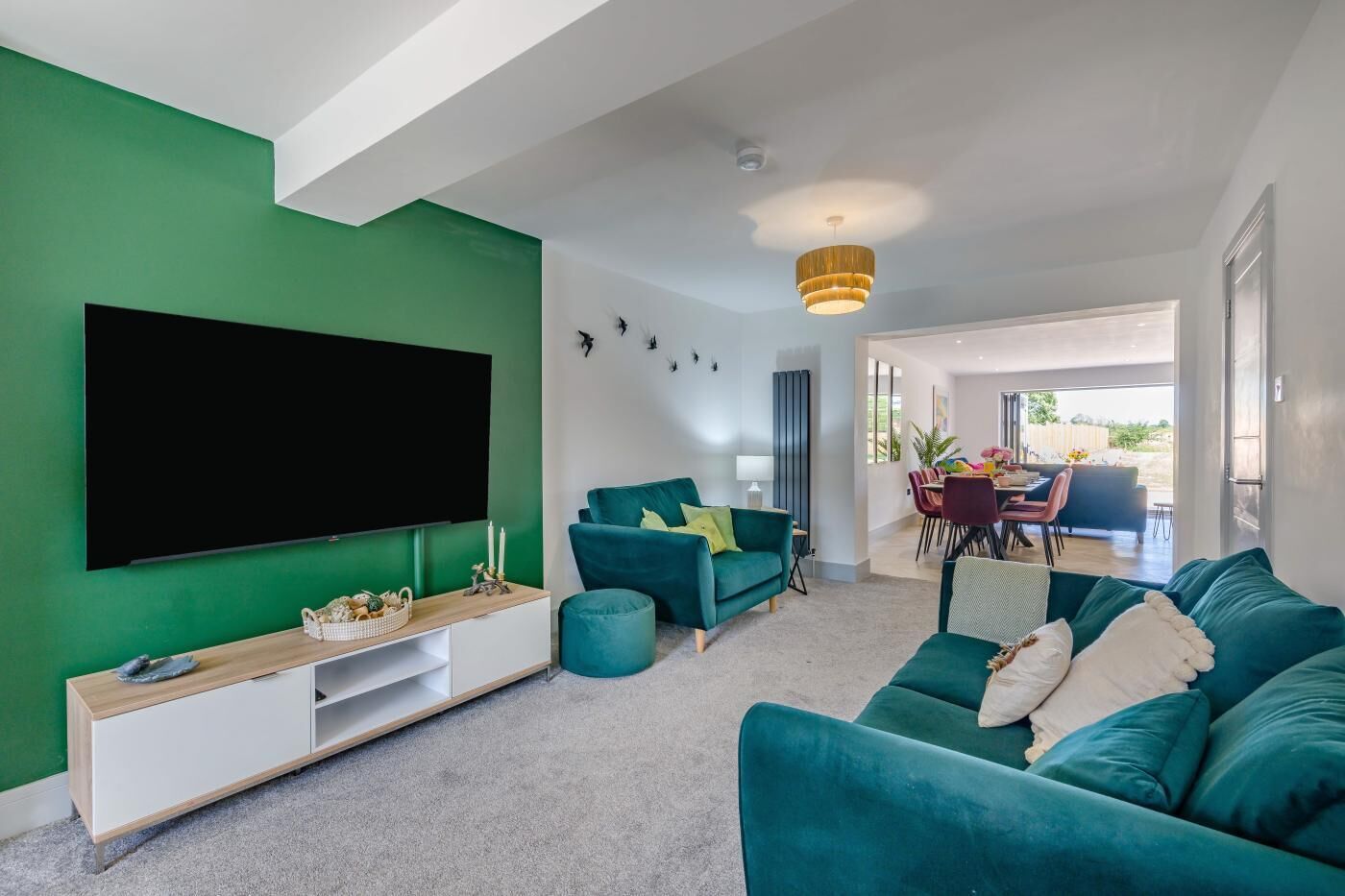 A modern living room with a large flat-screen TV, teal sofas, and a dining area with a table and chairs.