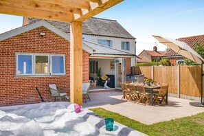Ein Garten mit einer Holz-Pergola, einem Patio-Tisch mit Stühlen, einem Ziegelhaus und einem eingezäunten Bereich.