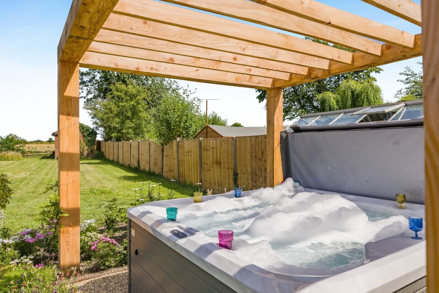 Un bain à remous sous une pergola en bois, situé dans un jardin avec une clôture en bois, une pelouse verdoyante et des arbres en arrière-plan.