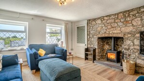 A cozy living room with a stone fireplace, a blue sofa set, and a view through large windows. - 3 Bed in Maudlam (oc-i32375) (Maudlam)