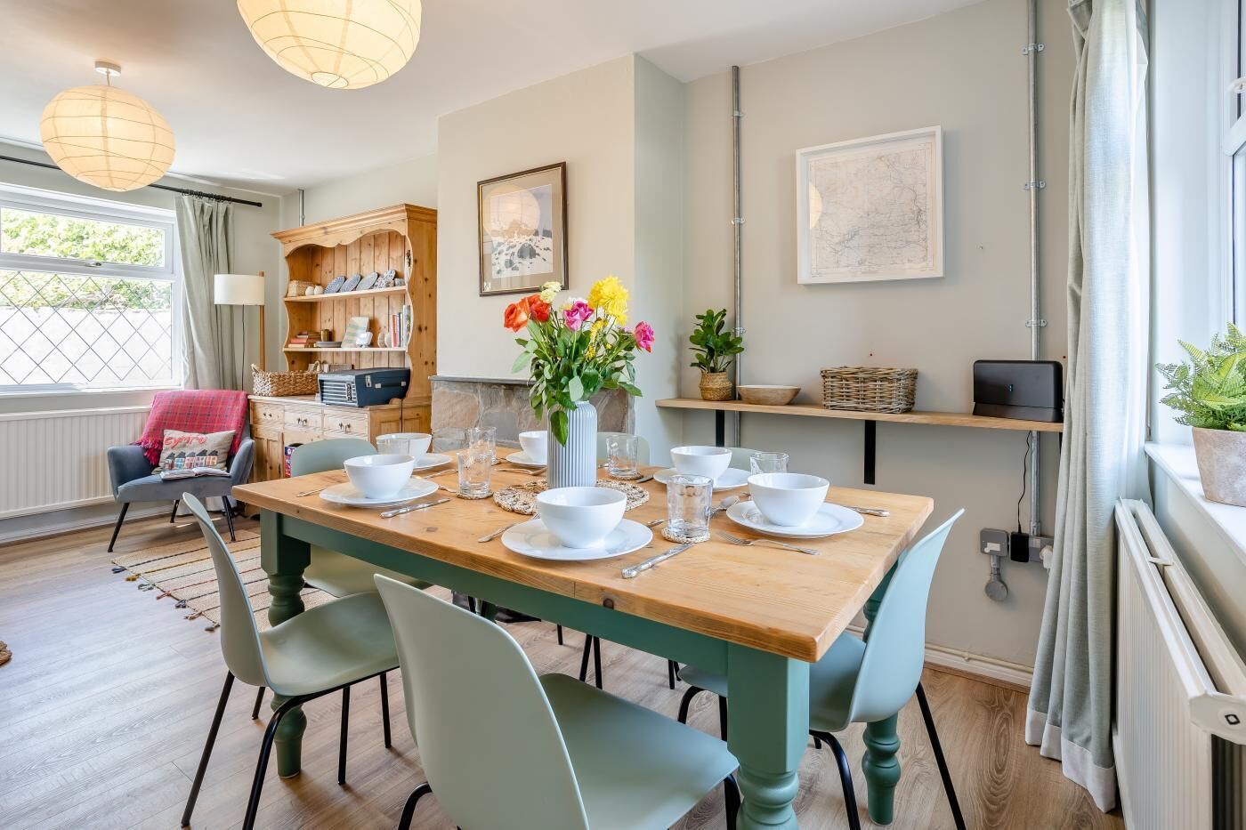 Un coin repas avec une table en bois dressée pour quatre personnes, entourée de chaises vert clair. On aperçoit un vase avec des fleurs sur la table, et une bibliothèque remplie de divers objets au fond.