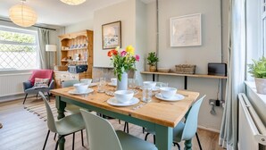 A dining area with a wooden table set for four, surrounded by light green chairs. There is a vase with flowers on the table, and a bookshelf with various items in the background. - 3 Bed in Maudlam (oc-i32375) (Maudlam)