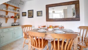 Ein Essbereich mit einem Holztisch, gedeckt für vier Personen, umgeben von Holzstühlen. Es gibt Regale mit Küchenutensilien und einen Spiegel über dem Tisch.