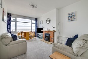 A living room with a fireplace, a dining area, and a large window with curtains. - 2 Bed in Southwold (oc-4east) (Southwold)