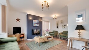 A modern living room with a flat-screen TV, green sofas, a wooden coffee table, and a chandelier. - 3 Bed in Whitby (oc-w015) (Whitby)