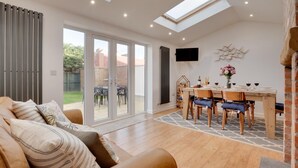 A modern living room with a sofa, a dining area with a table and chairs, a TV mounted on the wall, and a large sliding glass door leading to a garden. - 3 Bed in Whitby (oc-w015) (Whitby)