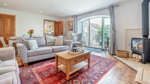 A spacious living room with a grey sofa, wooden coffee table, and a red patterned rug. A stone wall and a large window with curtains are visible. - 3 Bed in Cawton (oc-t32407) (Cawton)