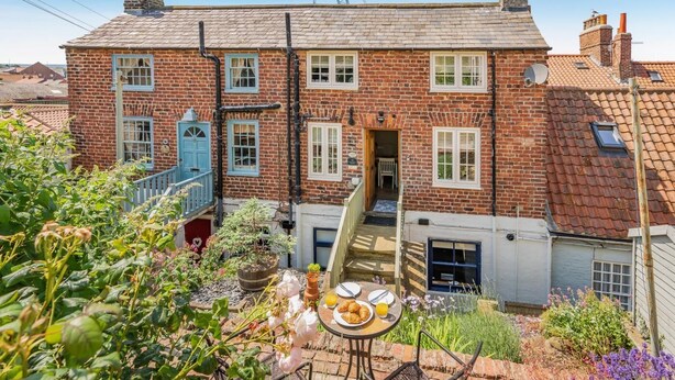 A brick house with a garden patio, a small table with plates and a chair, and a blue door. - 2 Bed in Whitby (oc-w904) (Whitby)
