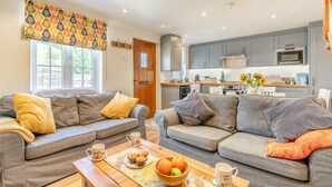 A living room with two grey sofas, a wooden coffee table with muffins and fruit, and a kitchen in the background. - 2 Bed in Whitby (oc-w904) (Whitby)