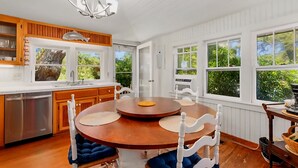 A kitchen with wooden cabinets, a stainless steel dishwasher, a round dining table with blue cushioned chairs, and large windows offering a view of greenery.