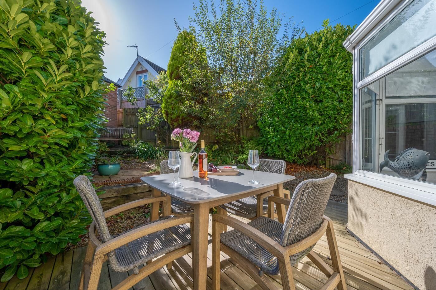 A wooden deck with a table and chairs, surrounded by greenery and a house in the background.