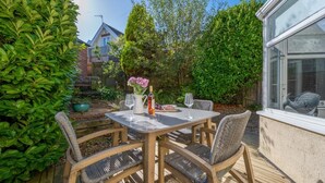 A wooden deck with a table and chairs, surrounded by greenery and a house in the background.