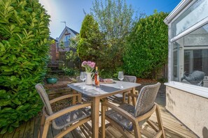A wooden deck with a table and chairs, surrounded by greenery and a house in the background.