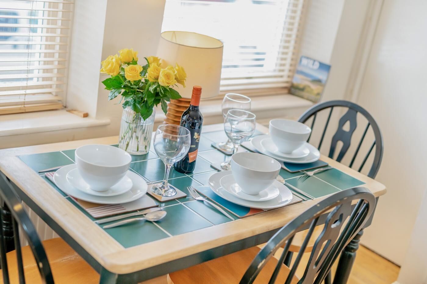 Une table à manger dressée avec des bols blancs, des verres à vin et une bouteille de vin, agrémentée d’un vase de fleurs jaunes et d’une lampe en arrière-plan.